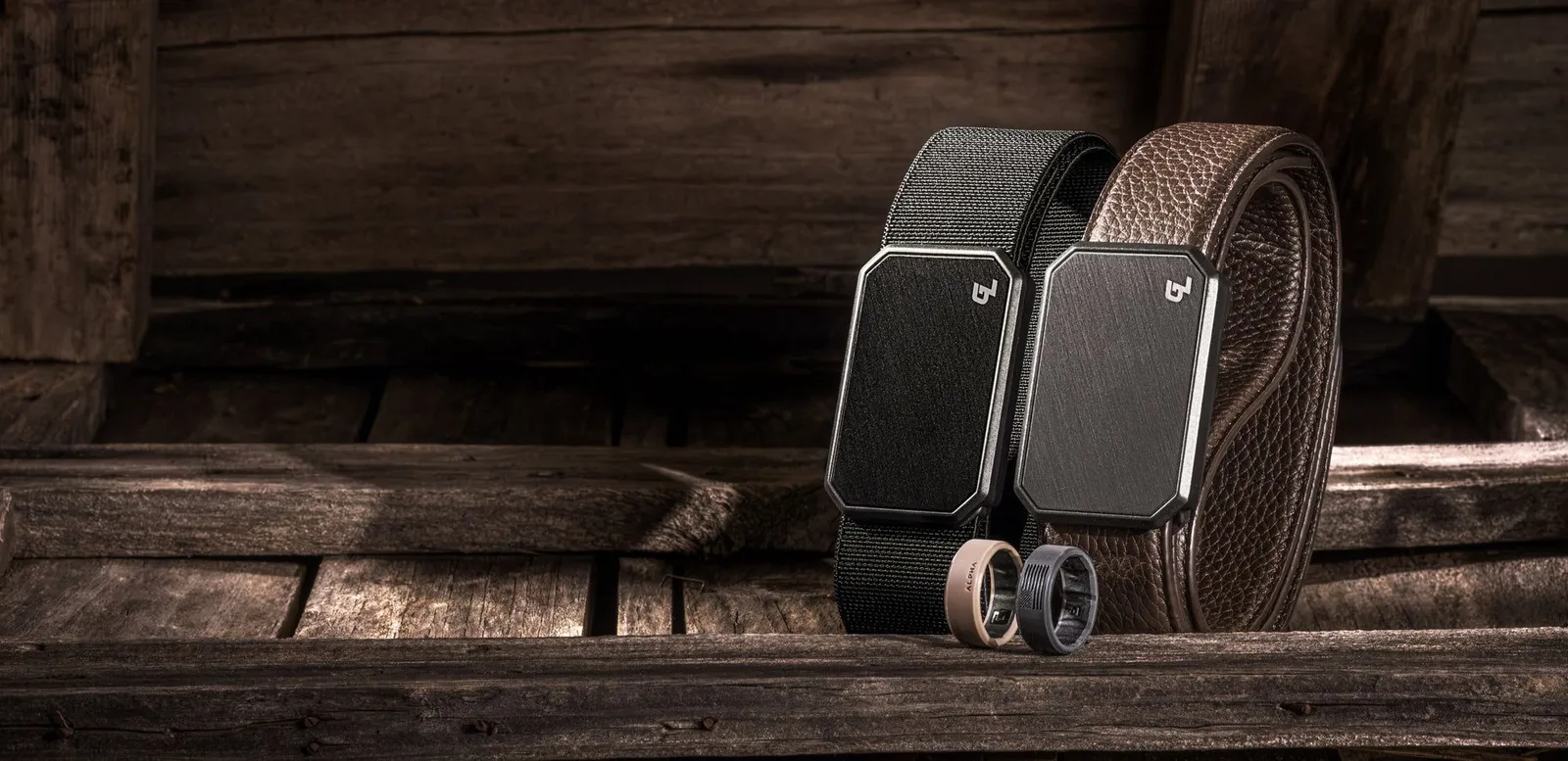 A black Groove Belt and a brown leather Groove Belt standing on weathered wood, buckles forward, with two Groove rings in front
