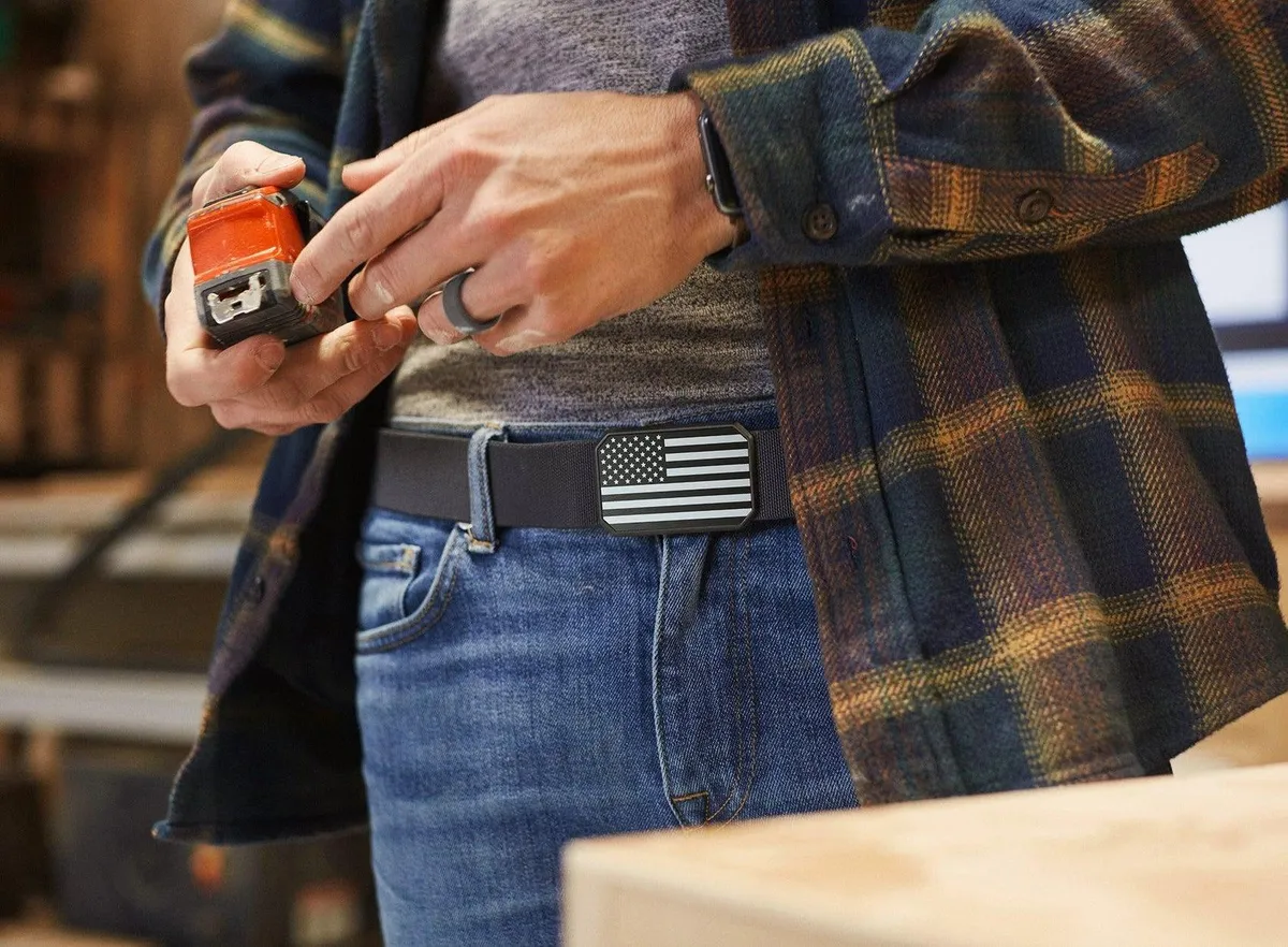 Person wearing a plaid jacket and jeans with an American flag belt buckle, holding a tool.
